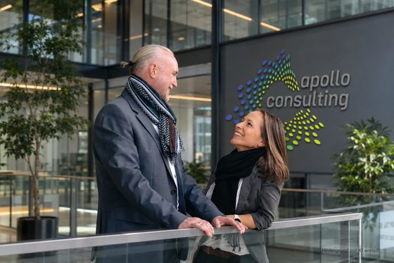 Volker Loibl und Melanie Reuß im Gespräch, im Hintergrund die Logo-Wand von apollo consulting